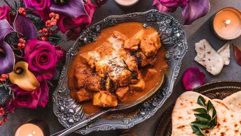 Closeup on a serving dish of paneer makhani. Pink and purple floral arrangements sit on the left and a plate of naan sits on the right. 