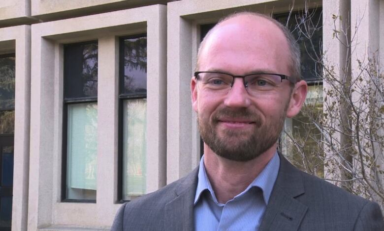  A man wearing glasses stands in front of a grey building.