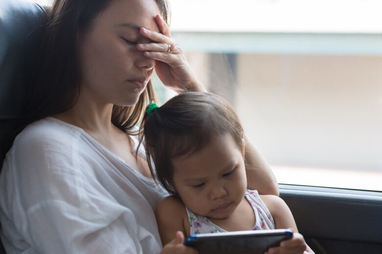 A mother holds her head in her hand while her child looks at an electronic screen.