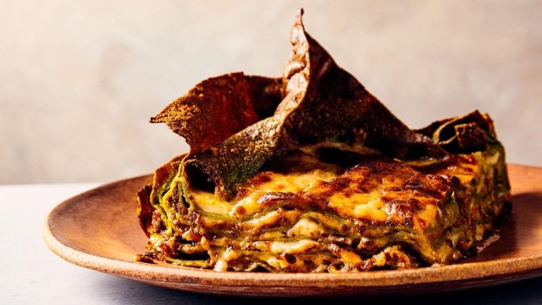 closeup on a large piece of cheesy lasagna. multiple layers of pasta are visible and the top layer is crispy. it's on a brown plate on a beige surface. 