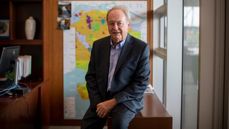 A man in a suit sits on a table in an office. A map of the city of Surrey is behind him.