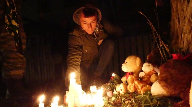 A man in a jacket leans forward with one hand out behind candles on a dark night. The candlelight shines on a pile of stuffed animals.