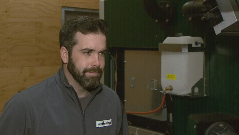 Man with dark brown hair and a beard stands next to a furnace in what looks like a basement. 