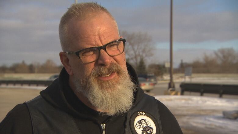 A man with a white beard squints a bit on a sunny winter day. He wears a hoodie with a badge that says 