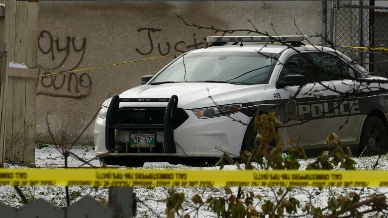 A police car sits behind yellow police tape, next to a stucco wall with grafitti on it.