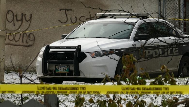 A police car sits behind yellow police tape, next to a stucco wall with grafitti on it.