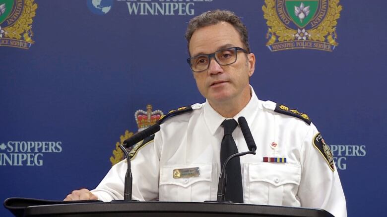 A man in a uniform shirt and tie stands behind a podium with microphones.