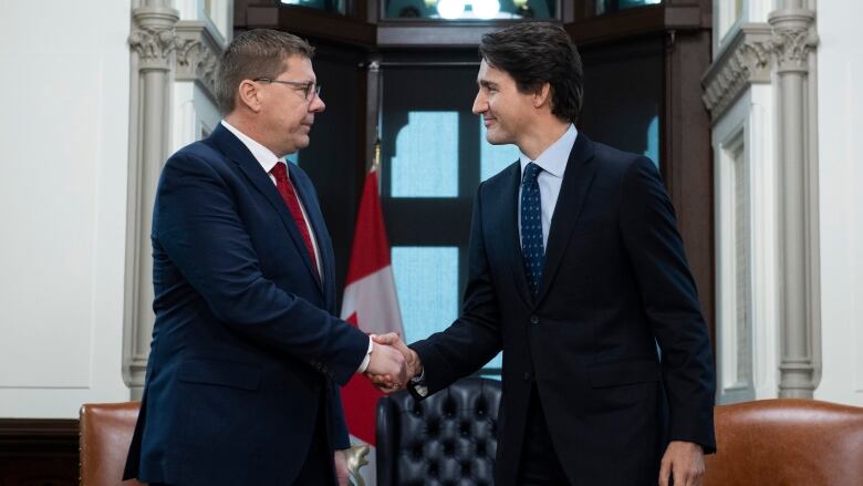 Two men wearing suits shake hands in front of a Canadian flag and a desk.