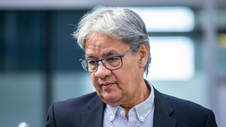 Ed John looks down. He is an Indigenous man with grey hair, wearing blue-coloured square glasses.