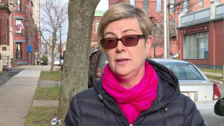 A woman standing outside by a car and a tree. She is wearing sunglasses, a pink scarf and a black jacket.