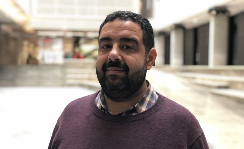 A person with a beard and mustache wears a sweater on top of a collared shirt. He has a slight smile.