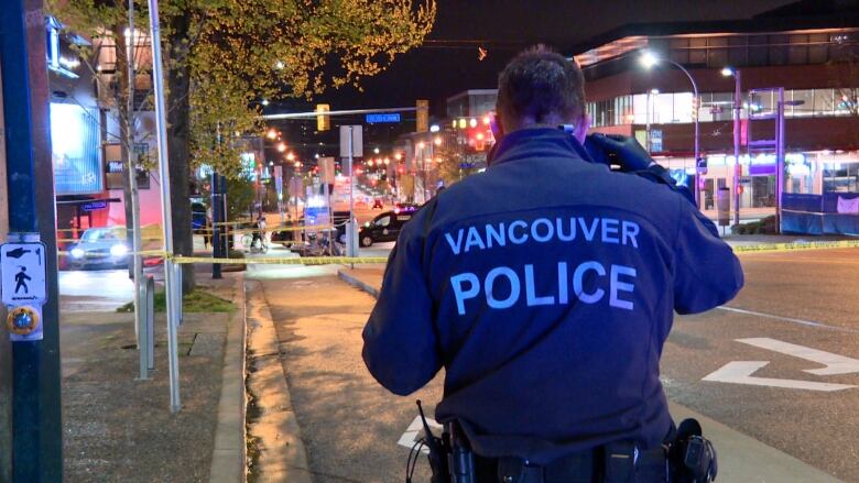 A man in uniform with the words Vancouver Police on the back and a utility belt on his side listens to a device near his ear while walking down a lit sidewalk at night.