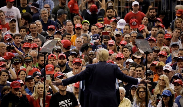 A man with his back to the camera spreads his arms grandly, facing a large crowd of spectators. 