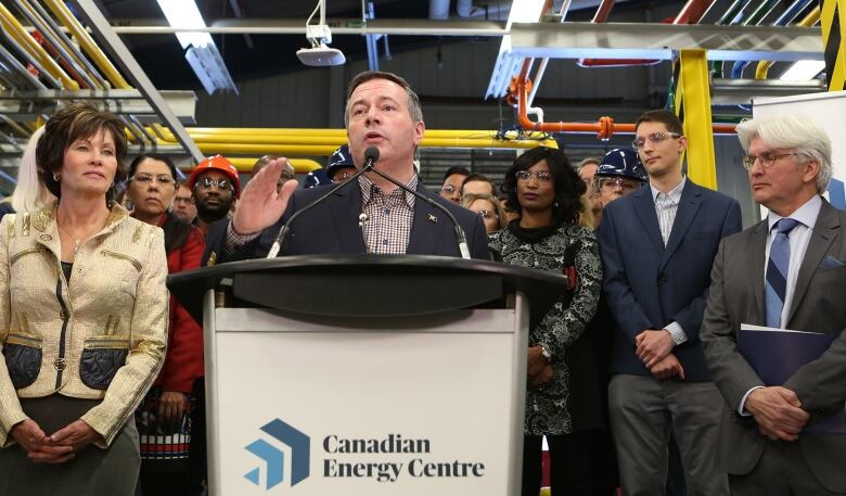 A man in a black suit stands at a podium. He is flanked by a large crowd of men and women in business attire. 