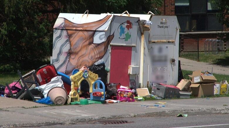 Piles of garbage including old toys and cardboard boxes surround to large metal donation bins 