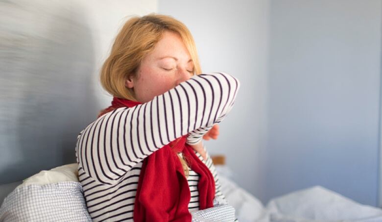 A blonde woman sits on a bed and coughs into her elbow. 