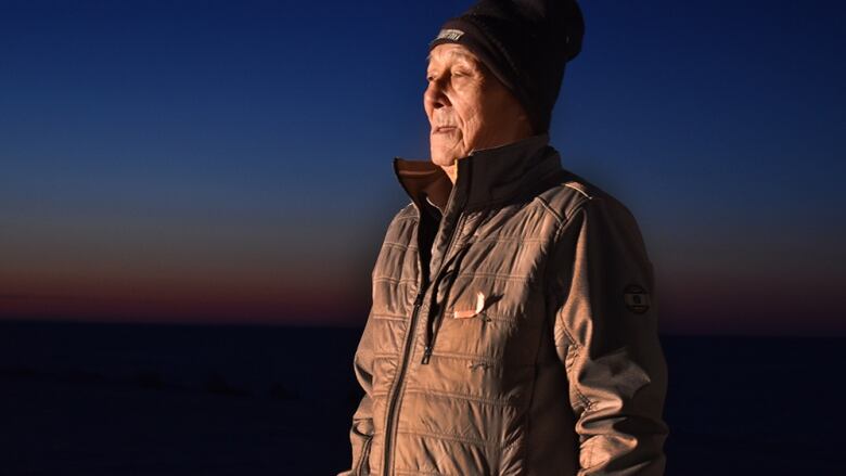 A man with a brown coat, brown mitts, and a black hat stares into light against a dark outdoor background.