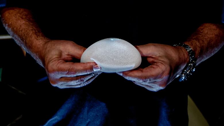 The hands of a plastic surgeon in blue scrubs hold a breast implant.