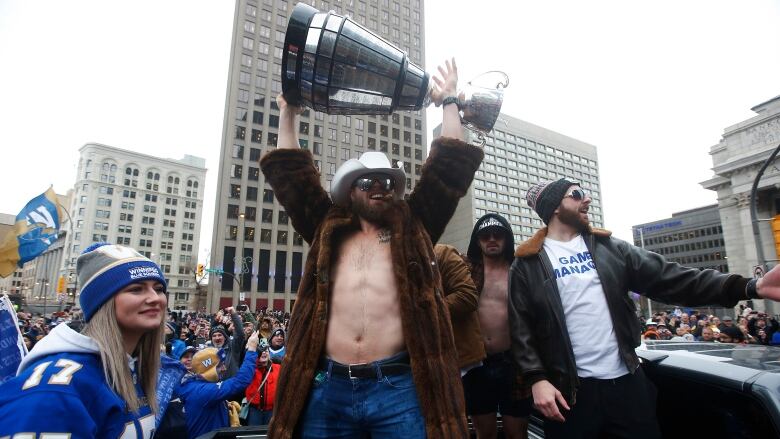 A man in fur coat and cowboy hat, with a bare chest under the open coat, holds up a trophy.