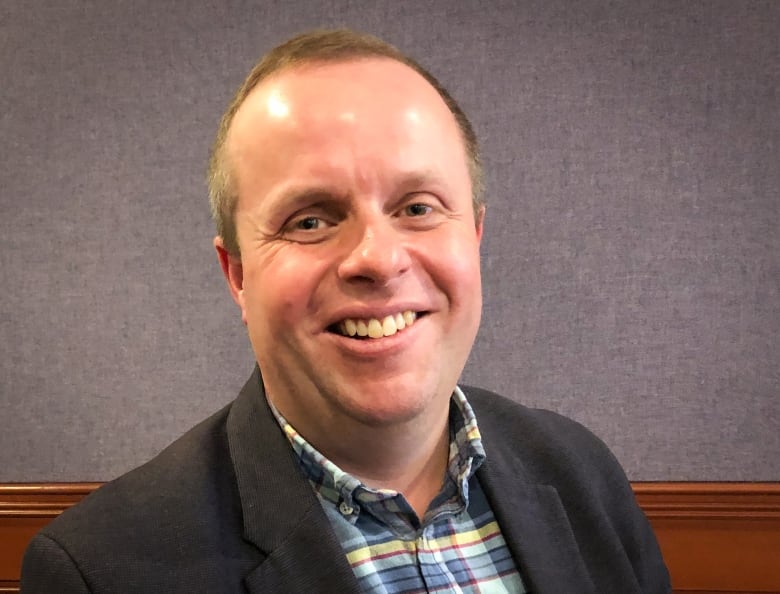 A man with light brown hair wearing a dark jacket and a plaid shirt smiles.