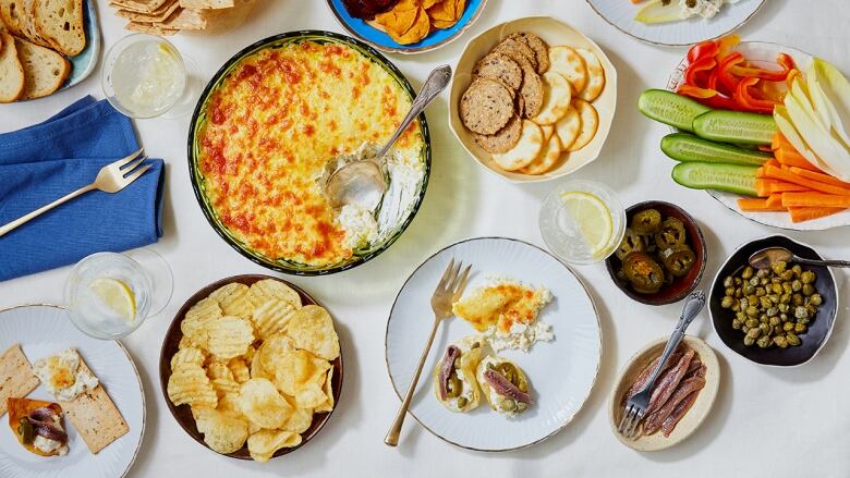 An overhead shot of a spread of dishes on a white table, featuring a dish of baked broccoli dip, crackers, breads and vegetables. 