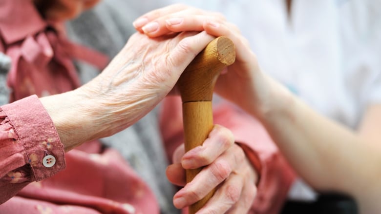 Hands of a senior rest on a cane, the hand of a caregiver on top of it.