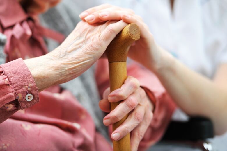 Hands of a senior rest on a cane, the hand of a caregiver on top of it.