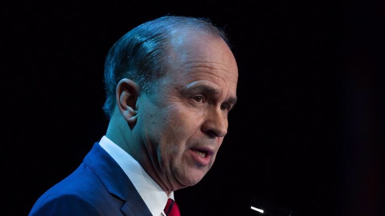 A man in a suit and red tie speaks at a microphone.