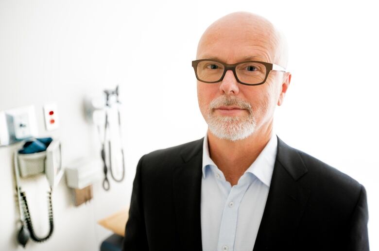 Bearded man with glasses in a business suit, standing in a medical office. 