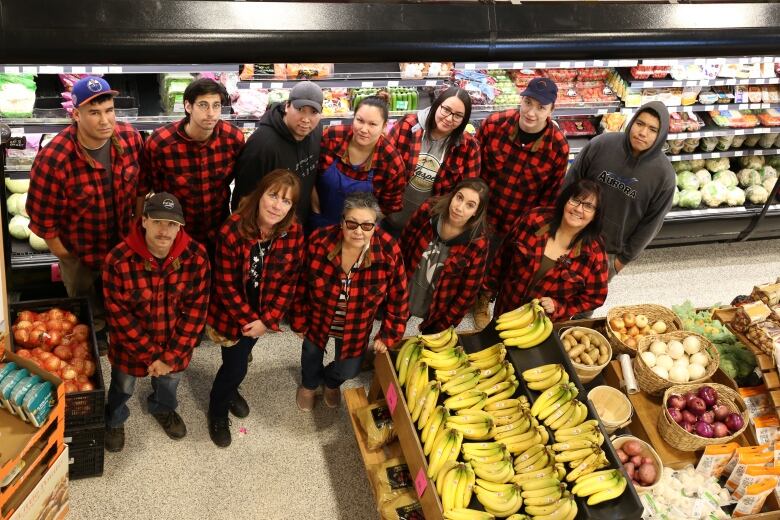 A group of workers in red and black plaid jackets stand in a grocery store, looking up to the camera. 