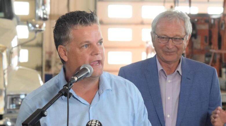 One man with short dark grey hair speaks animatedly into a microphone wearing a button up dress shirt that is sky blue. Behind him, to the right of the photo, stands a man with short grey hair and glasses, smiling and wearing a dark blue suit jacket over a lighter dress shirt.