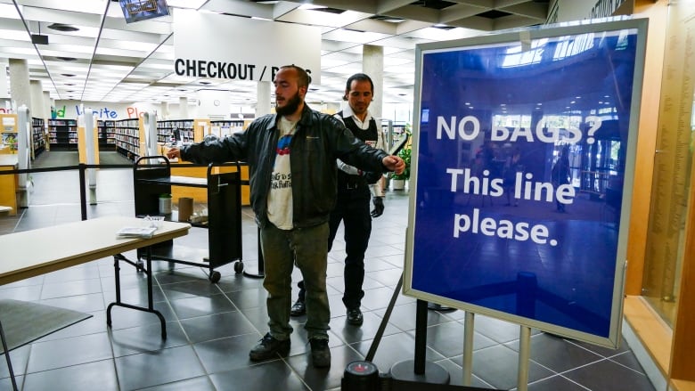 A man is standing at the entrance to a library with his arms out stretched while a security guard holds a metal detector over him.