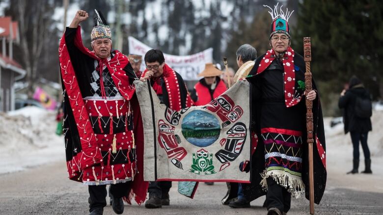 Two men in traditional clothing walk, holding a banner between them. The man on the left holds his right fist in the air, while the man on the right holds a staff in his left hand.