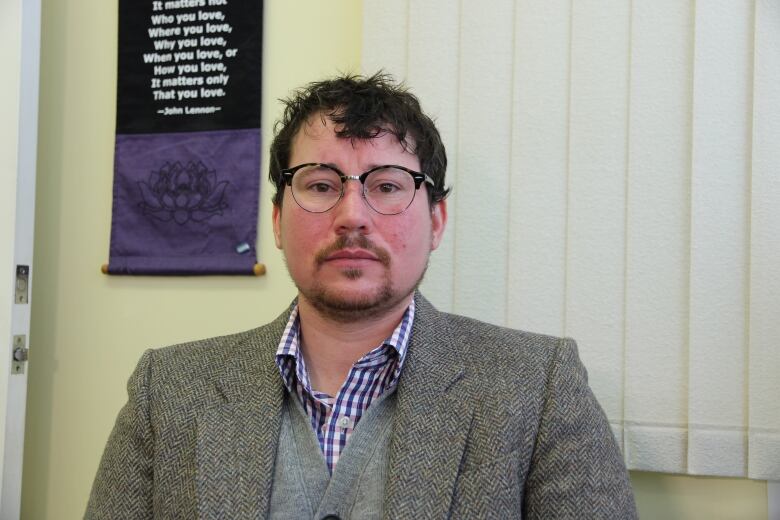 Man with brown hair and moustache wearing glasses and a grey tweed jacket and grey woolen vest looks directly into camera.