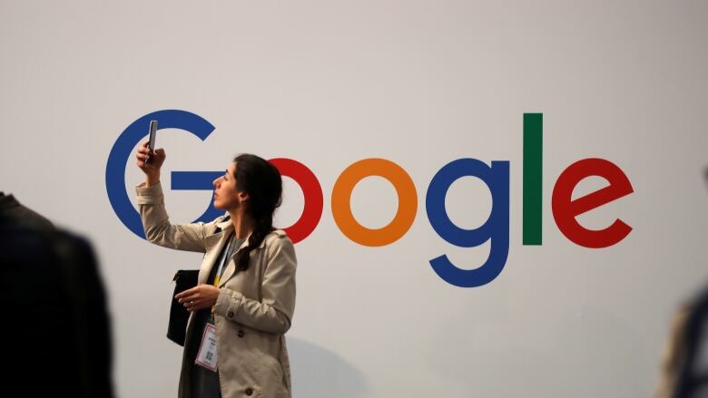 A woman standing in front of a wall bearing the multicoloured Google logo lifts her phone up.