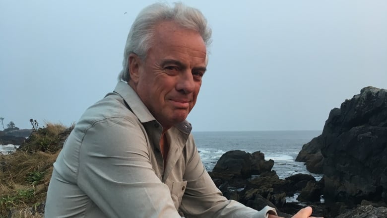 A man sits outside, on rocks beside a body of water