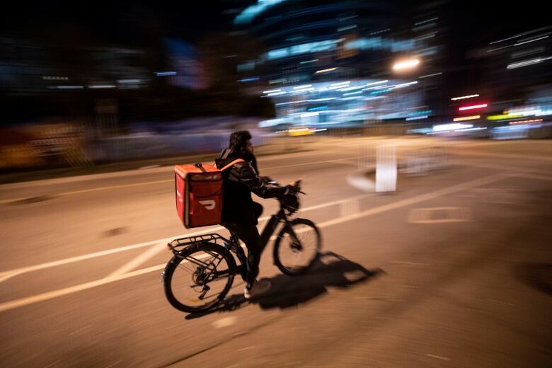 A cyclist with an orange DoorDash delivery bag bikes at night down a Vancouver street.