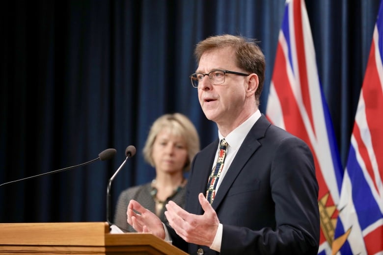 B.C. Health Minister Adrian Dix stands in front of a podium while Dr. Bonnie Henry stands in the blurry background.