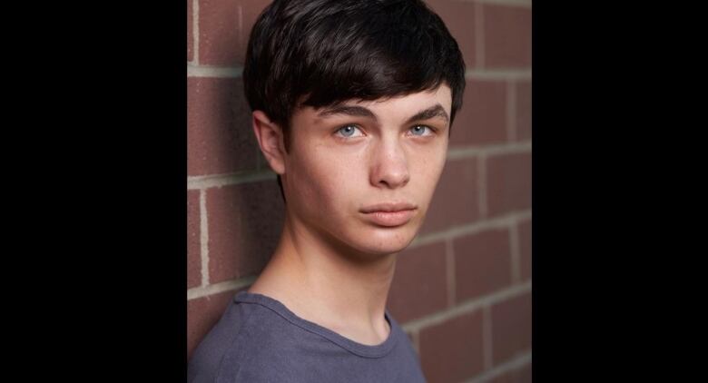 A young man with black hair and blue eyes, dressed in a grey-blue t-shirt is pictured in front of a brick wall, gazing intently at the camera.