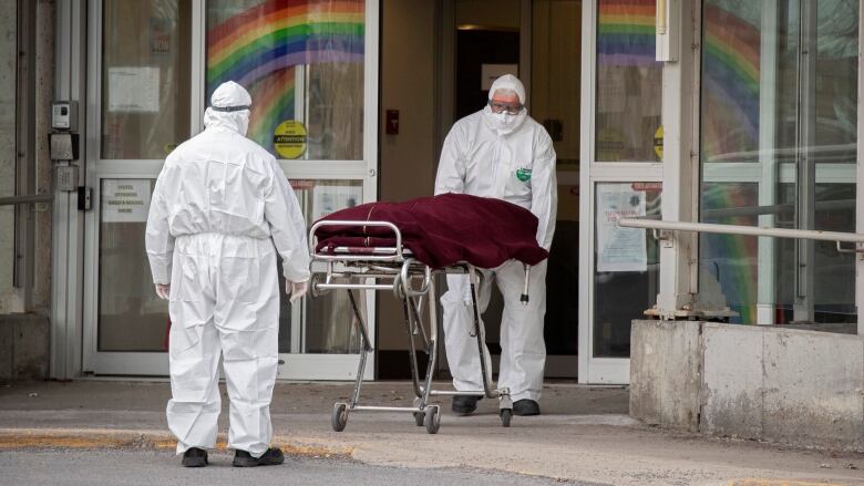 Two medics wearing PPE transport a covered body on a gurney inside the hospital. 