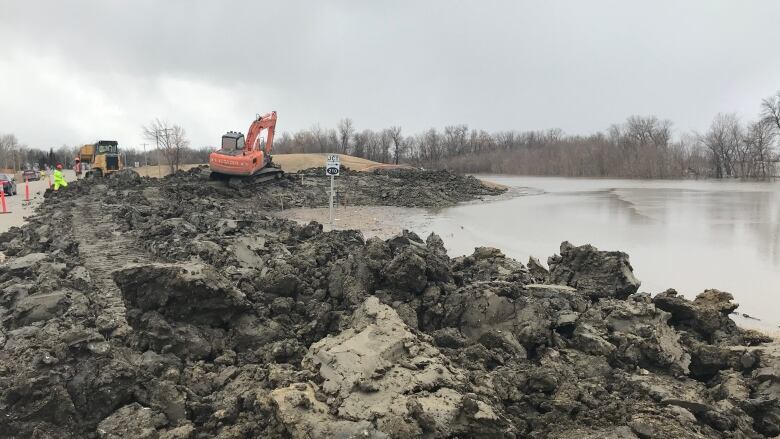 Mounds of dirt can be seen in the foreground, with some water to the right side of the frame. In the distance, excavators and construction workers wearing high-visibility yellow clothing can be seen working.