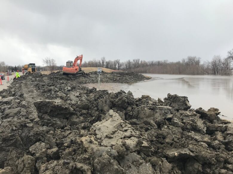 Mounds of dirt can be seen in the foreground, with some water to the right side of the frame. In the distance, excavators and construction workers wearing high-visibility yellow clothing can be seen working.