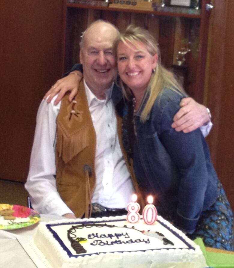 An elderly man in a leather vest smiles for the camera. His has his arm around a blonde woman in a blue shirt who stands to his left. 