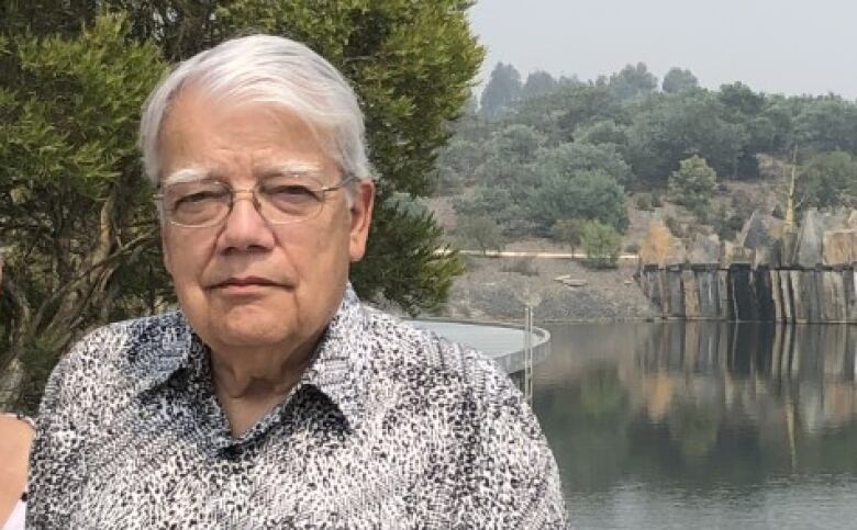 A man with white hair and glasses poses for a photo outside.