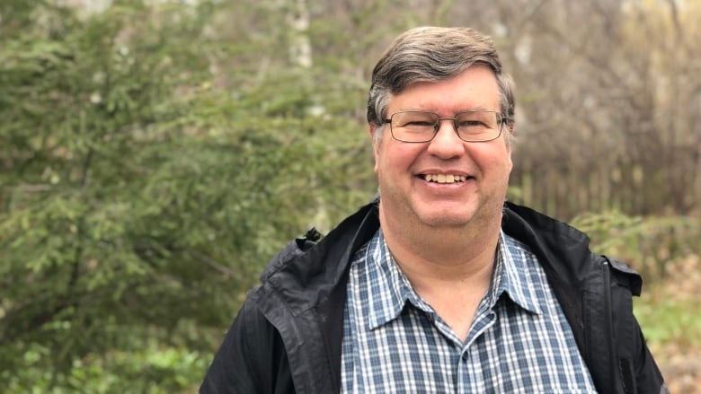 A man with grey hair and glasses smiles outside in front of some trees.