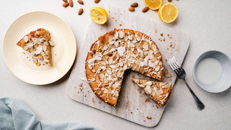 Overhead shot of a cake topped with almond slices and drizzled with white icing. It's sitting on a white marble cutting board on a white table. 2 slices are cut from the cake and one slice is sitting on a white plate next to it. 