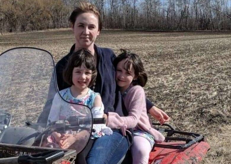 A woman sits with two young, smiling girls on a red ATV in a farm field.