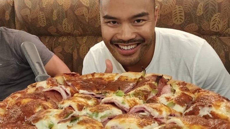 A man sitting in a restaurant booth, smiling towards the camera from behind a large pizza.