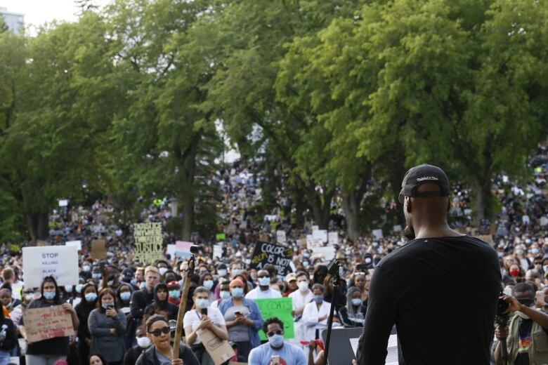 A Black man addresses a crowd of people, some holding protest signs. 