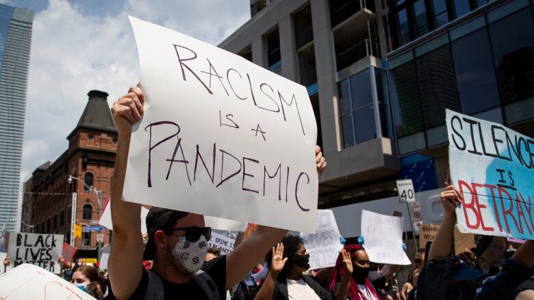 A crowd is seen with people holding signs that say things such as 'Racism is a pandemic' and 'Silence is betrayal.'